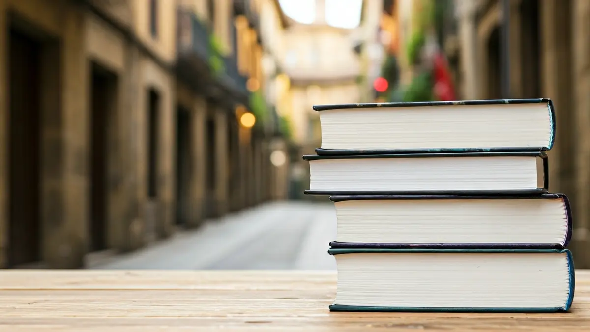 Liburu pila bat egurrezko mahai baten gainean, atzealdean Euskal Herriko hiri zahar bateko kale bihurgunetsu bat lausotuta.