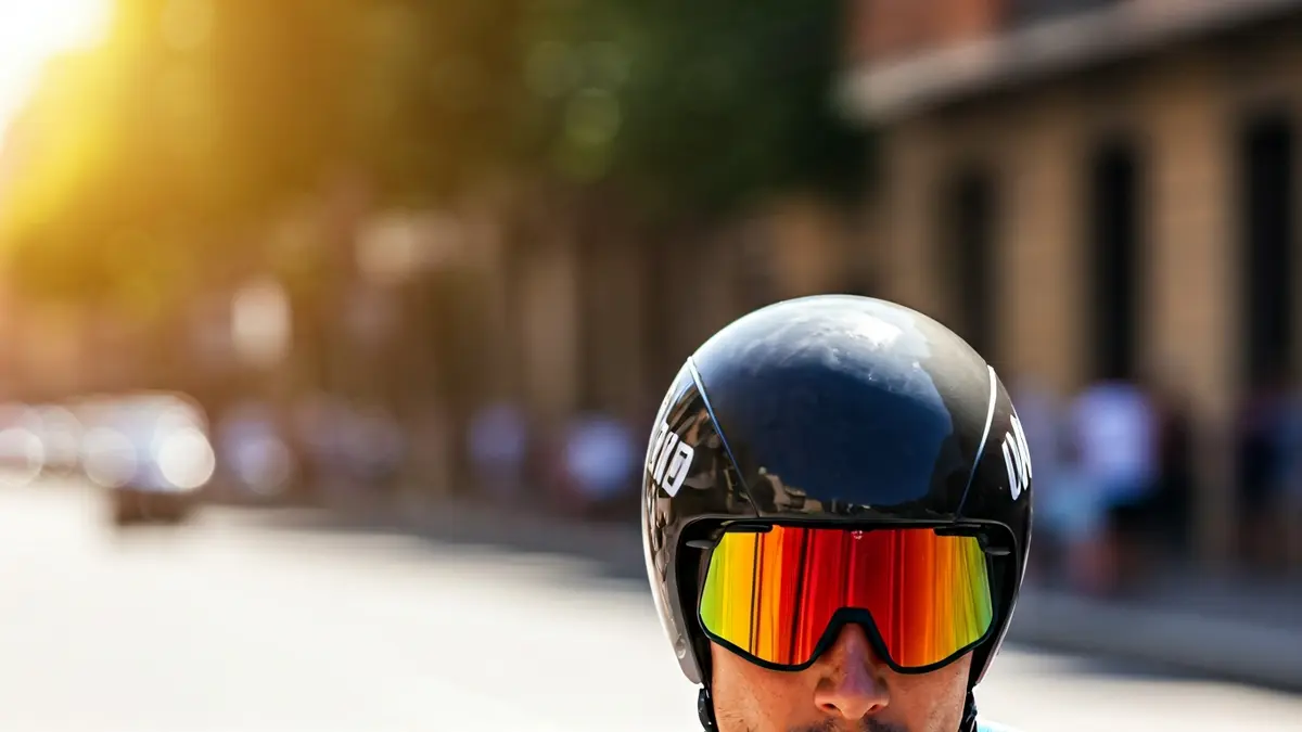 Txirrindulari baten kasko aerodinamikoaren eta bisore ilunaren hurbileko irudia, atzealde lausoan Bilboko hiri-paisaia eguzkitsu eta beroa ikusten dela, erlojupeko batean, abiadura eta beroa azpimarratuz.