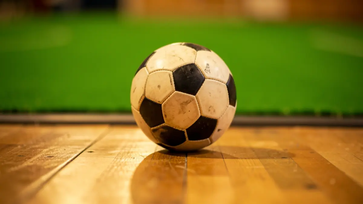 Close-up of a futsal ball on a wooden court floor, with blurred green astroturf in the background.