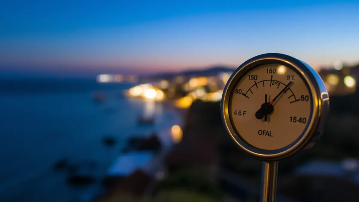 A thermometer showing a high temperature reading, with a blurred coastal landscape of the Basque Country in the background under a clear night sky.