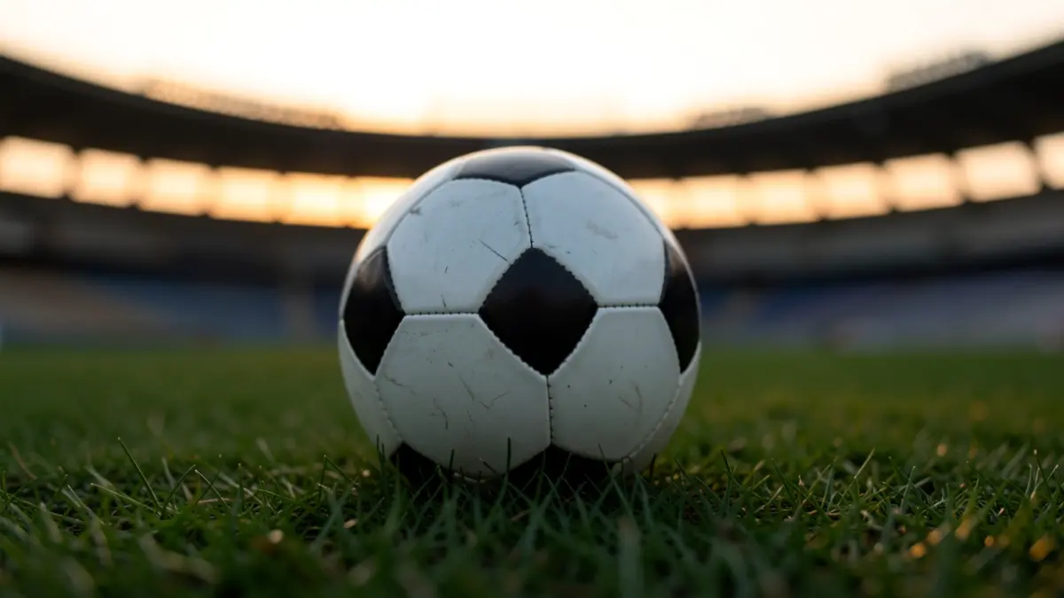 Un balón de fútbol sobre hierba, con la silueta difuminada de un estadio al fondo.