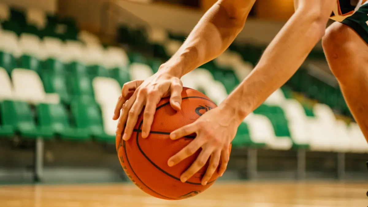Imagen genérica: Manos de un jugador de baloncesto botando un balón, fondo de estadio difuminado.