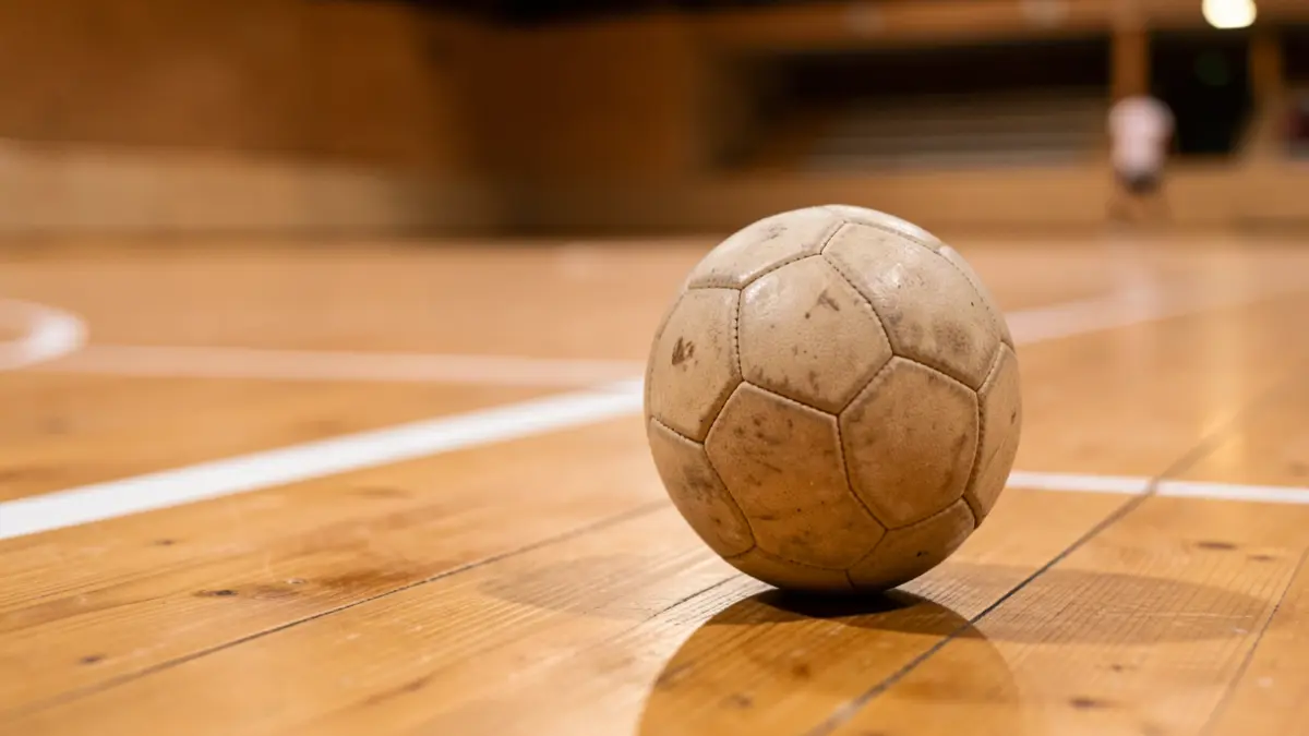 Close-up of a pelota ball resting on the wooden floor of a fronton court, white lines of the court blurred in the background, warm indoor lighting, shallow depth of field.