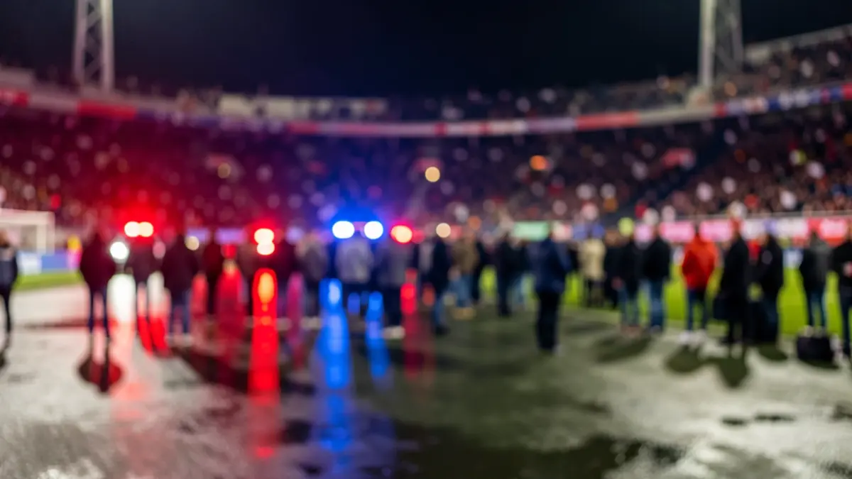 Generic image: football stadium crowd at night, with red and blue emergency lights reflecting on wet ground, shallow depth of field, no identifiable people or vehicles.