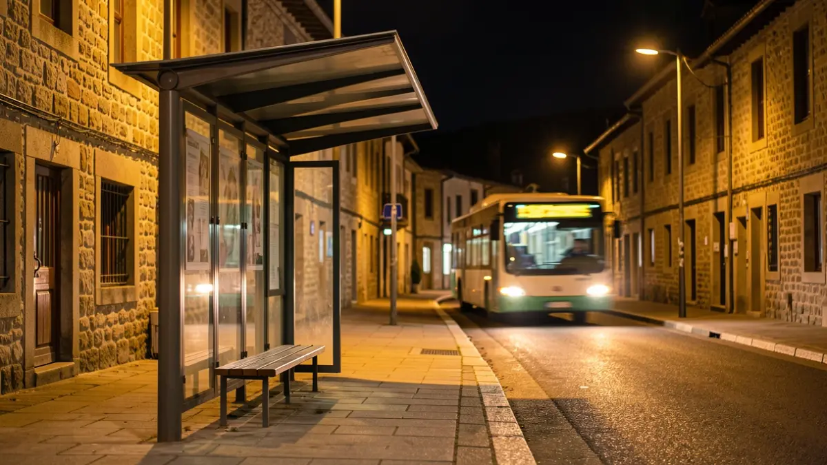 Una parada de autobús nocturna con luces artificiales y un autobús acercándose a lo lejos.