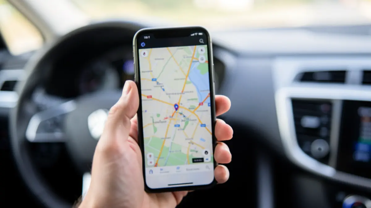 Primer plano desenfocado de la pantalla de un teléfono móvil mostrando un mapa GPS, sostenido por una mano parcialmente visible. Al fondo, el salpicadero y el volante de un coche fuera de foco.