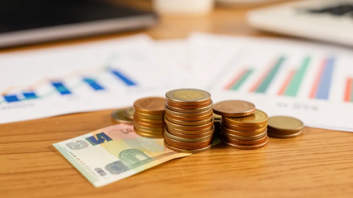 Generic image: A stack of euro coins and banknotes on a wooden desk, with blurred graphs in the background.